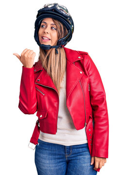 Young beautiful woman wearing motorcycle helmet smiling with happy face looking and pointing to the side with thumb up.