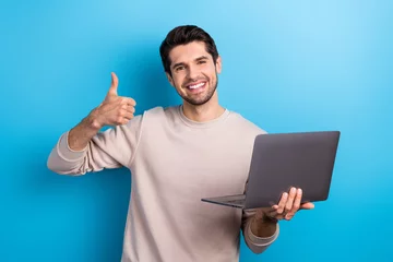 Tuinposter Portrait of young nice guy hold laptop show thumb up wear beige sweatshirt isolated on blue color background © deagreez