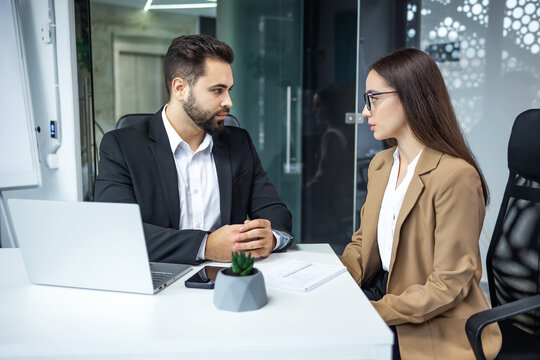 Two caucasian business partners dressed in formal clothes sitting together at office desk and talking. Charming dark haired woman attentively listening bearded man sharing ideas of new project.