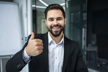 Bearded positive man looks at the camera and happily shows a thumbs up gesture.