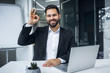 Smiling businessman showing ok sign, like gesture, in modern office building. Man in a formal suit is looking at the camera. Template for advertising. - 775083675
