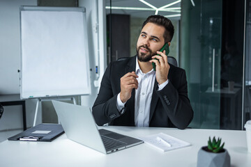 Handsome businessman using a computer, talking on the mobile phone while working in office - 775084255