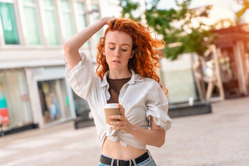 unhappy young redhead woman in jeans and white shirt drinking take away coffee on street. lifestyle concept