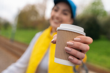 happy young woman in yellow  waistcoat and blue cap drinking take away coffee, tea on the street...