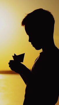 Young boy is making a paper ship standing near the river at the evening sunset. Silhouette of the boy playing with paper boat outside Vertical video