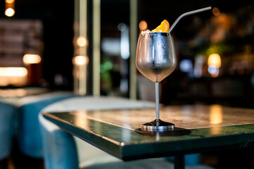 A chilled cocktail in a metal glass, adorned with an orange slice, rests on a bar table in a dimly...