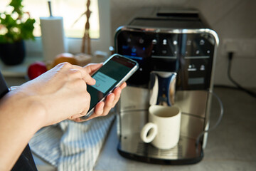 Person uses smartphone app to operate coffee machine. Woman preparing fresh cup of coffee with...