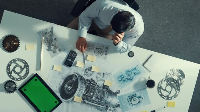 Top view of engineer looking at phone while working on tablet with green screen at meeting table with turbine engine or jet engine sketch design and various metal gear scatter around. Alimentation.