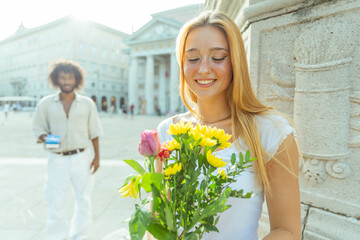 Happy woman with a bouquet of flowers, her partner behind with a box with the ring is about to propose