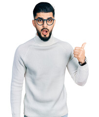 Naklejka premium Young arab man with beard wearing elegant turtleneck sweater and glasses surprised pointing with hand finger to the side, open mouth amazed expression.