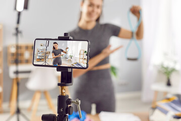 Sporty slim young woman instructor or fitness blogger in sportswear recording video for blog about online training using phone camera on tripod talking about sport exercises with rubber band