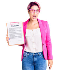 Young beautiful woman with pink hair holding clipboard with contract document winking looking at the camera with sexy expression, cheerful and happy face.