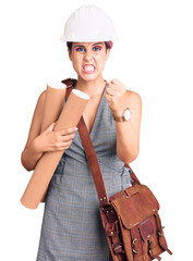 Young beautiful woman wearing architect hardhat and leather bag holding blueprints annoyed and frustrated shouting with anger, yelling crazy with anger and hand raised