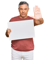 Handsome middle age man holding blank empty banner with open hand doing stop sign with serious and confident expression, defense gesture