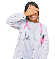Beautiful hispanic woman wearing doctor uniform and stethoscope smiling and laughing with hand on face covering eyes for surprise. blind concept.