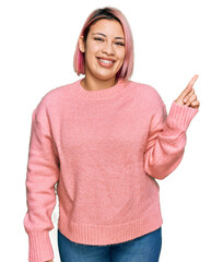 Hispanic woman with pink hair wearing casual winter sweater with a big smile on face, pointing with hand finger to the side looking at the camera.