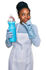 Young african american woman wearing apron holding detergent bottle serious face thinking about question with hand on chin, thoughtful about confusing idea