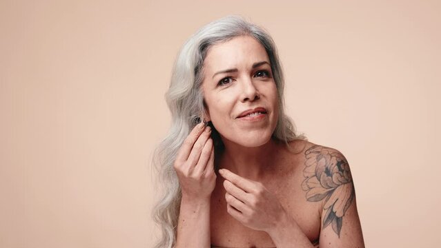 A senior woman with grey hair stands poised as she adjusts her earring, her tattoo visible, embodying timeless beauty and confidence.