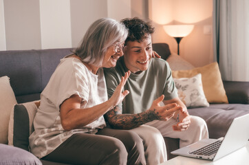 Video call concept. Happy senior retired woman sitting on sofa with young nephew waving hands video calling by laptop using technology for online webcam connection with distant family or friends - obrazy, fototapety, plakaty