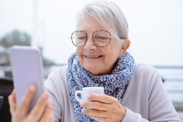 Portrait of smiling white haired senior woman sitting outdoors at cafe table looking at social apps on smart phone enjoying a break with an espresso coffee