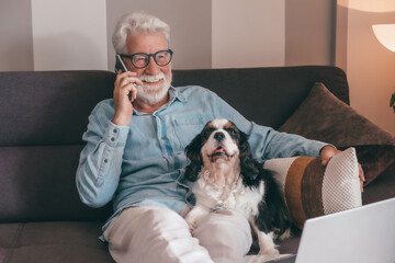 Senior man talking on mobile phone sitting on sofa with his cavalier king charles dog. Elderly male...