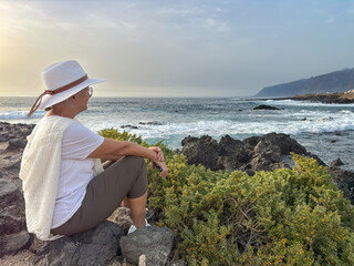 Rear view of senior woman sitting on a rocky beach watching ocean waves crashing with white foam....