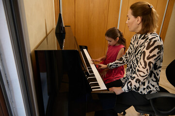 Confident authentic female pianist, music teacher giving an individual piano lesson to her little...