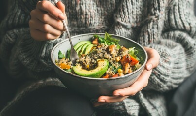 Woman in a grey sweater eating a healthy salad bowl with avocado, quinoa and vegetables 