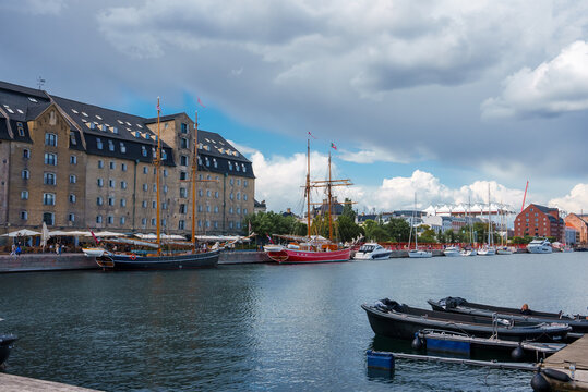 Copenhagen waterfront scene with calm waters, moored boats, and a tall ship. Historical yellow and red buildings under a cloudy blue sky create a serene, picturesque setting.