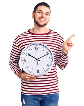 Young handsome man holding big clock smiling happy pointing with hand and finger to the side