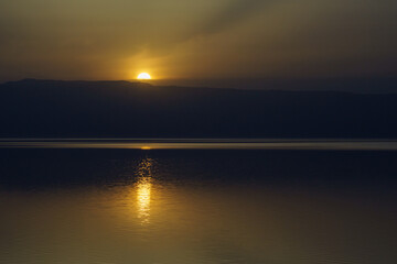 Jordan. Dead Sea. Calm at sea. Sunset on Dead Sea coast. Orange sun is reflected in water. There is light path in water from reflection of sun. Opposite bank with mountains is Israeli territory.
