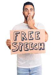 Young hispanic man holding free speech banner covering mouth with hand, shocked and afraid for...