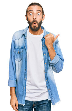 Attractive man with long hair and beard wearing casual denim jacket surprised pointing with hand finger to the side, open mouth amazed expression.