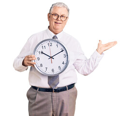 Senior grey-haired man wearing business clothes and holding clock celebrating victory with happy smile and winner expression with raised hands