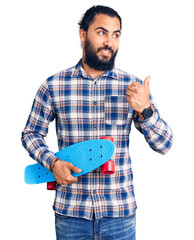 Young arab man holding skate pointing thumb up to the side smiling happy with open mouth