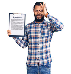 Young arab man holding clipboard with agreement document smiling happy doing ok sign with hand on eye looking through fingers