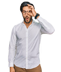 Young hispanic man wearing business shirt and glasses doing ok gesture shocked with surprised face, eye looking through fingers. unbelieving expression.