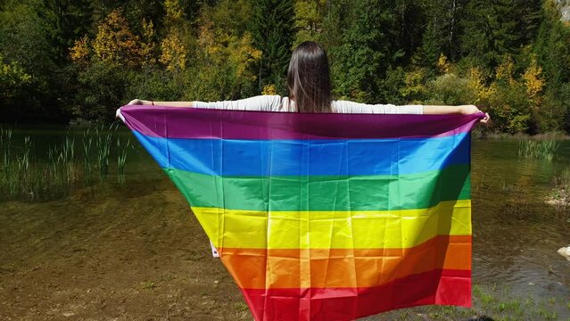 Pride month LGBTQ. Young female waving LGBT gender identity flag against beautiful lake and nature. Freedom concept 