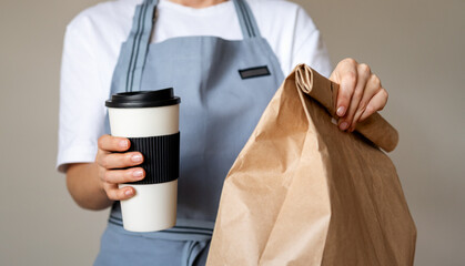 Takeaway meal in hands of waitress. Travel coffee mug and brown paper package bag with food inside. 