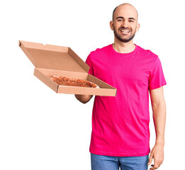 Young handsome man holding delivery pizza cardboard box looking positive and happy standing and smiling with a confident smile showing teeth
