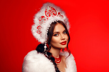 Beautiful brunette fashion woman portrait in fur coat and kokoshnik (woman's headdress in old Russia). Pretty model girl with red lips and plait hairstyle isolated on red studio background.