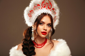 Beautiful brunette woman winter portrait in fur coat and kokoshnik (woman's headdress in old Russia). Pretty model girl with red lips and plait hairstyle isolated on grey studio background.