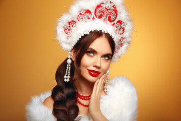 Beautiful brunette fashion woman portrait in fur coat and kokoshnik (woman's headdress in old Russia). Pretty model girl with red lips and plait hairstyle isolated on yellow studio background.