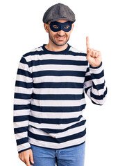 Young handsome man wearing burglar mask showing and pointing up with finger number one while smiling confident and happy.