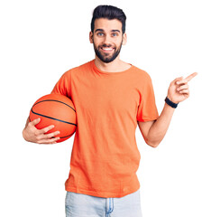 Young handsome man with beard hoilding basketball ball smiling happy pointing with hand and finger...
