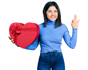 Young brunette woman with blue eyes holding valentine gift doing ok sign with fingers, smiling friendly gesturing excellent symbol