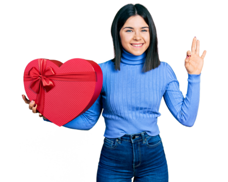 Young brunette woman with blue eyes holding valentine gift doing ok sign with fingers, smiling friendly gesturing excellent symbol