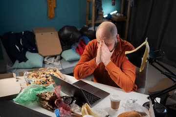 Depressed man working or studying at home sitting in a dirty room in piles of garbage and leftover...