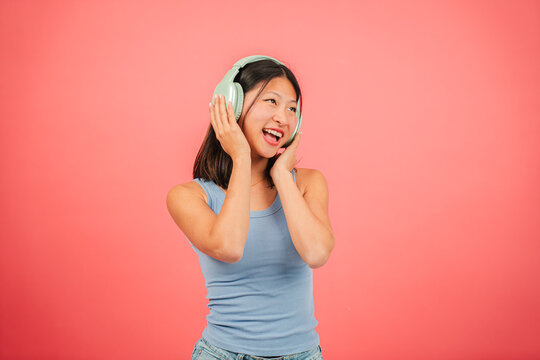 Young asian woman dancing music using a wireless headphone and a streaming cellphone app at red backdrop. Joyful chinese teenage female listening a song with earphones at pink color background wall