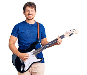 Young handsome man with curly hair playing electric guitar looking positive and happy standing and smiling with a confident smile showing teeth